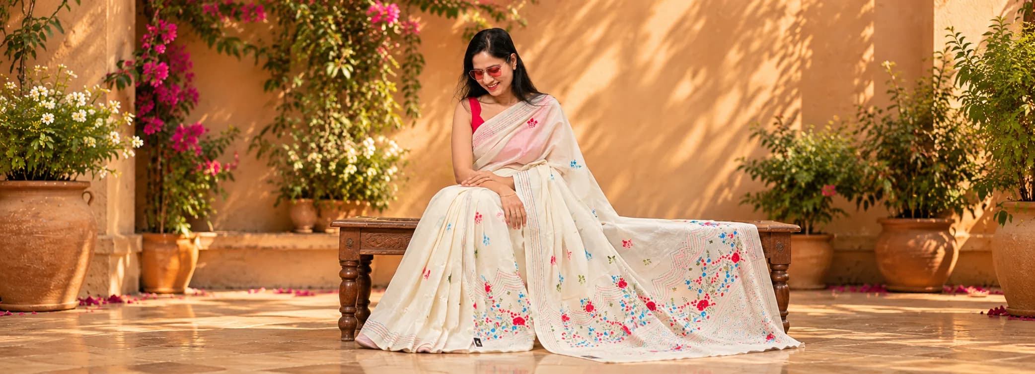 A smiling model in a white saree with floral embroidery, seated on a bench beneath bougainvillea