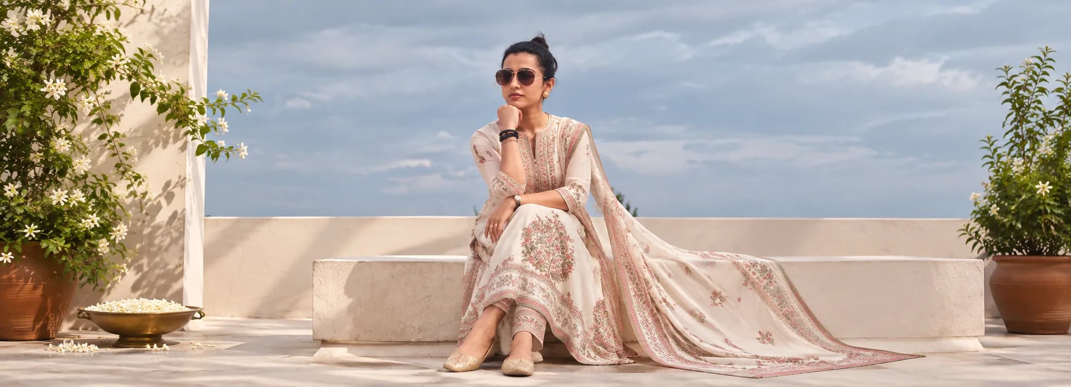 A model in a cream printed kurta set with a dupatta, seated on a white stone ledge under a soft blue sky